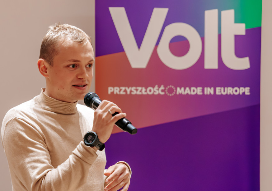 Przeowdniczący Maciej Tadeusiak podczas debaty. W ręku trzyma mikrofon; przemawia. W tle banner z napisem VOLT. Przyszłość made in Europe.