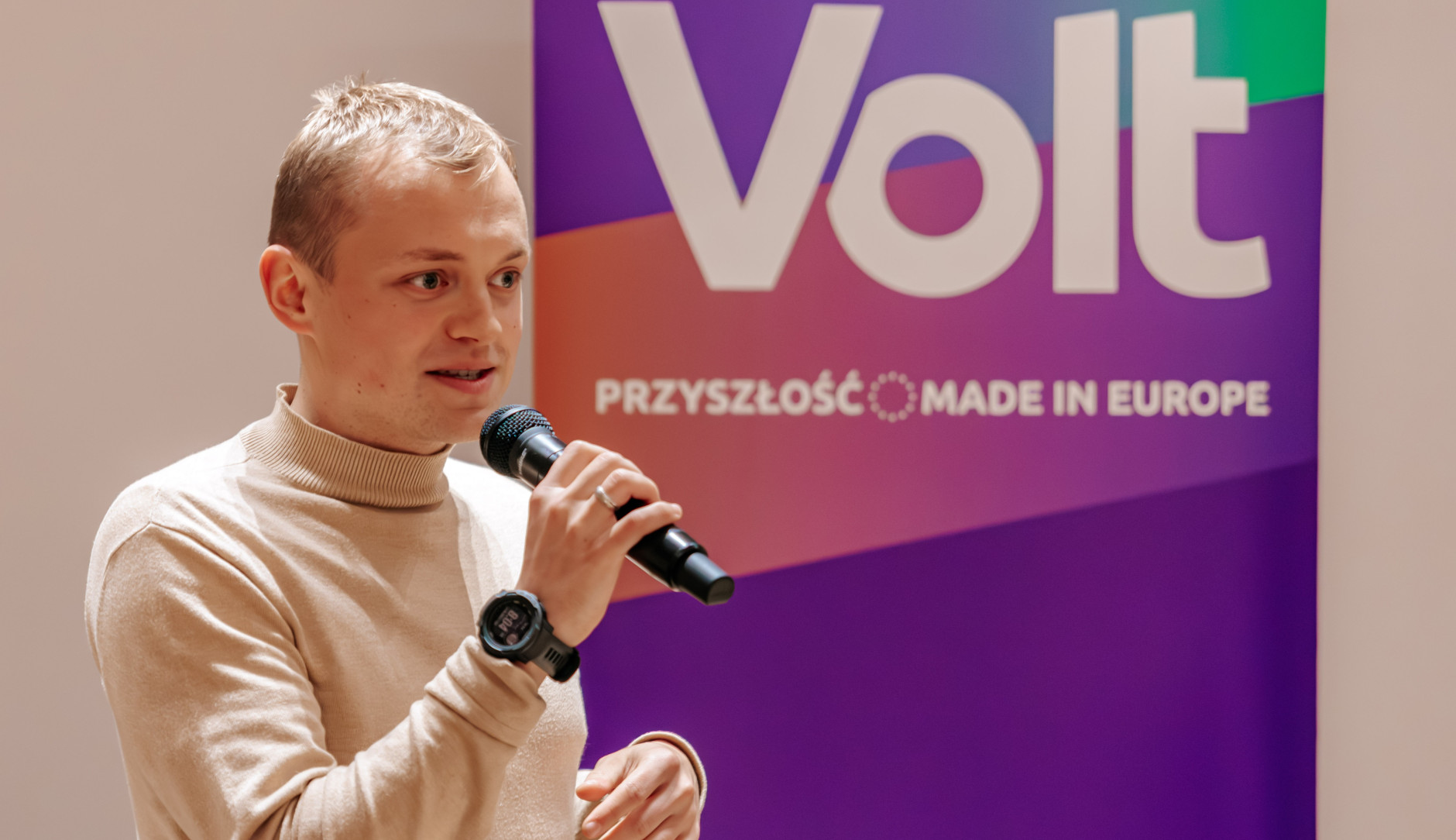 Przeowdniczący Maciej Tadeusiak podczas debaty. W ręku trzyma mikrofon; przemawia. W tle banner z napisem VOLT. Przyszłość made in Europe.