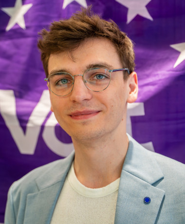 członek zarządu Karol Schubert na tle fioletowej flagi volt z 12 gwiazdami na wzór flagi europejskiej